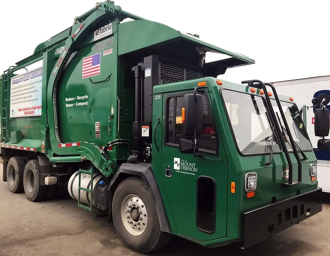 Green garbage truck parked with an American flag decal.